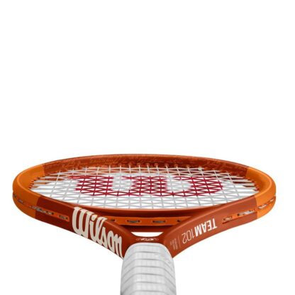 Throat view of orange tennis racquet with white grip. White Roland Garros logo at the bottom of the throat. Wilson in gold writing at the left side of the throat, and Team 102 on the right side. Wilson Roland Garros Team 102